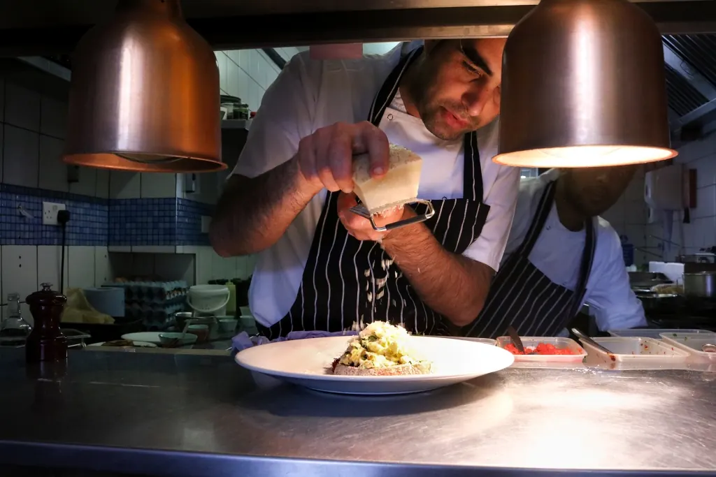 A chef at the pass at Manuka Kitchen grating parmesan over a plate under copper heat lamps